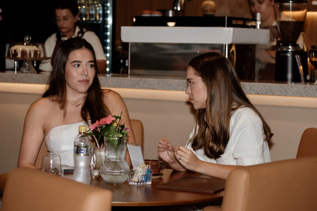 Duas clientes conversando à mesa, com o balcão da Casa ao fundo